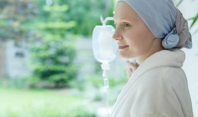 Eine junge Frau, welche Krebspatientin ist und ein Kopftuch trägt. Neben ihr steht ein Ständer mit einer Infusion und sie schaut in einen Garten. 