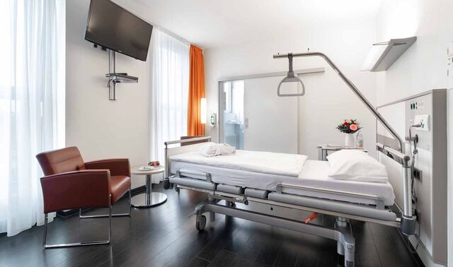 Patientenzimmer im Johanna-Etienne-Krankenhaus. In diesem befindet sich ein Bett, ein Fernseher und ein Tisch mit zwei Sesseln.