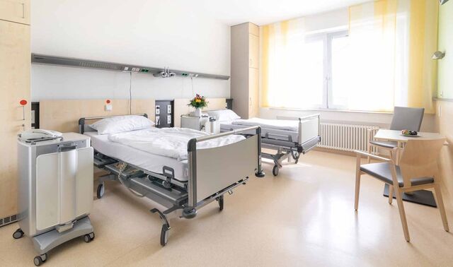 Patientenzimmer im Johanna-Etienne-Krankenhaus. In diesem stehen zwei Patientenbetten sowie ein Tisch mit zwei Stühlen. 