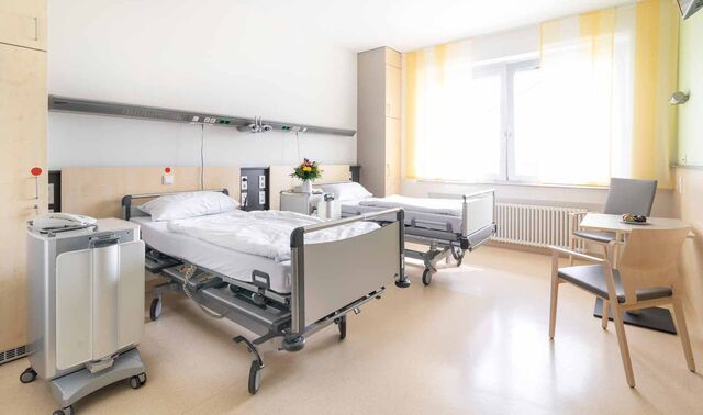 Patientenzimmer im Johanna-Etienne-Krankenhaus. In diesem stehen zwei Patientenbetten sowie ein Tisch mit zwei Stühlen. 