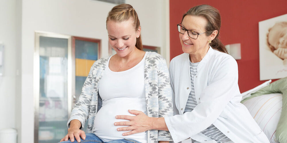 Eine Ärztin legt einer schwangeren Patientin eine Hand auf den Bauch. Beide lächeln und sitzen auf dem Patientenbett. 