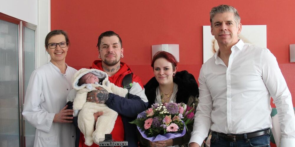 Vater und Mutter mit ihrem Neugeborenen schauen stehend in die Kamera. Links neben ihnen steht eine Ärztin, rechts neben ihnen ein Mann. Vor ihnen befindet sich Kameraequipment. 