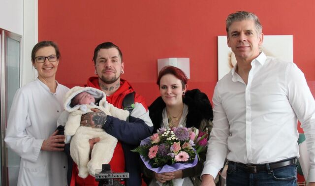 Vater und Mutter mit ihrem Neugeborenen schauen stehend in die Kamera. Links neben ihnen steht eine Ärztin, rechts neben ihnen ein Mann. Vor ihnen befindet sich Kameraequipment. 