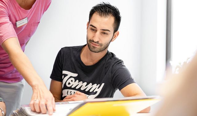 Ein junger Mann sitzt vor einer Akte an einem Tisch. Eine Frau, die neben ihm steht zeigt auf etwas in der Akte.