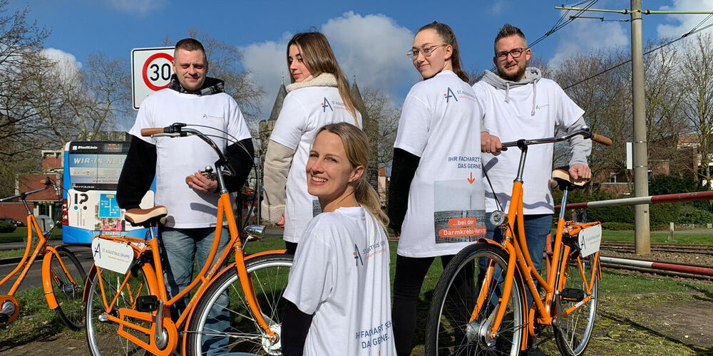 Einige junge Menschen tragen Augustinus Tshirts und haben zwei Fahrräder bei sich. Eine junge Frau kniet vor den anderen vier. 