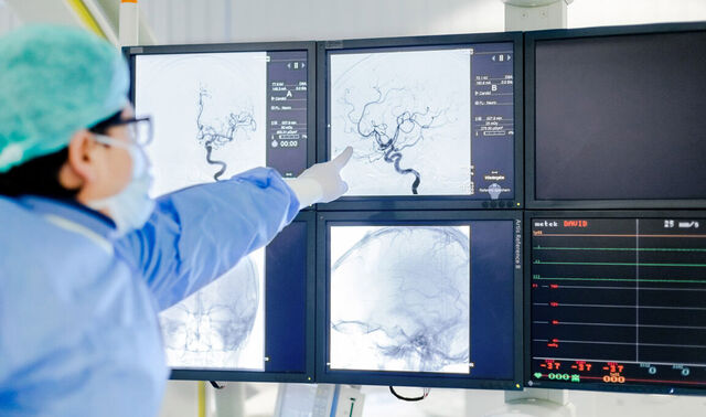 Eine Person in Operationskleidung zeigt auf einen von mehreren Bildschirmen, auf welchem kleine Äderchen sind. 