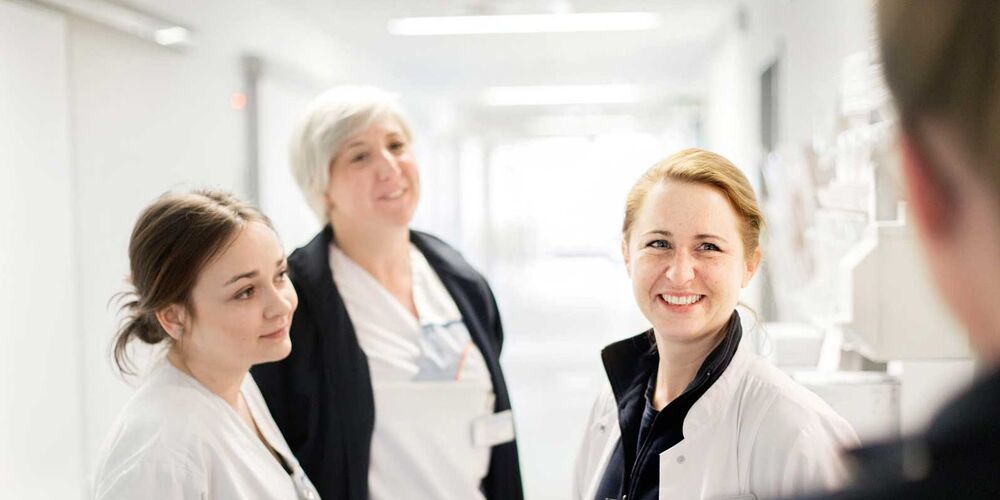 Drei Frauen stehen Flur eines Krankenhauses einer anderen Person gegenüber. Zwei der Frauen tragen die Kleidung einer Krankenpflegerin, die dritte Frau trägt einen weißen Kittel. 