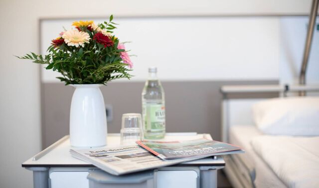 Tisch in einem Patientenzimmer, welcher neben einem Patientenbett steht. Auf diesem Tisch steht ein Blumenstrauße, eine Flasche Wasser sowie ein Glas und eine Zeitung und Zeitschrift.
