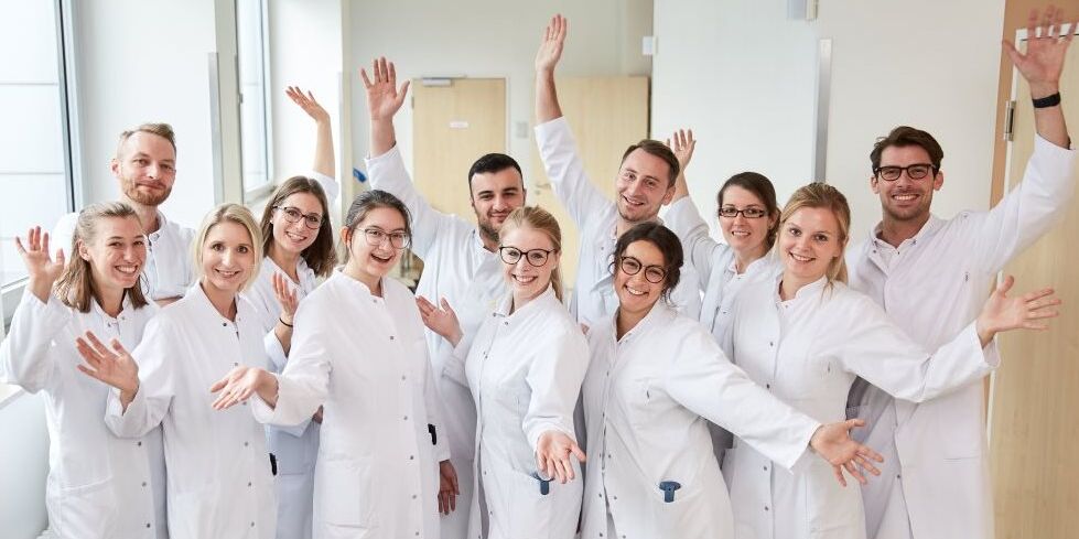 Teambild junger Ärzte und Ärztinnen, die in die Kamera schauen und lächeln und ihre Arme freudig nach oben gestreckt haben.