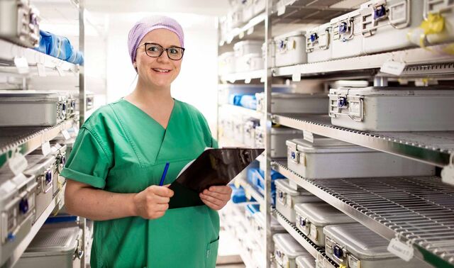 Porträt einer Mitarbeiterin in Operationskleidung zwischen zwei Regalen mit medizinischen Materialien.