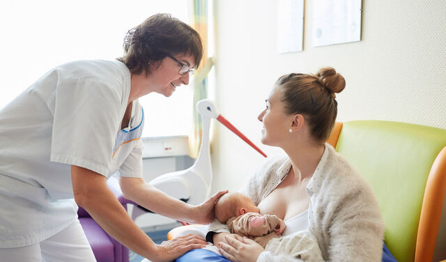 Eine junge Mutter sitzt und stillt ihr Neugeborenes hierbei steht eine Krankenpflegerin vor ihr und streichelt den Kopf des Säuglings. Die beiden Frauen schauen sich lächelnd an.