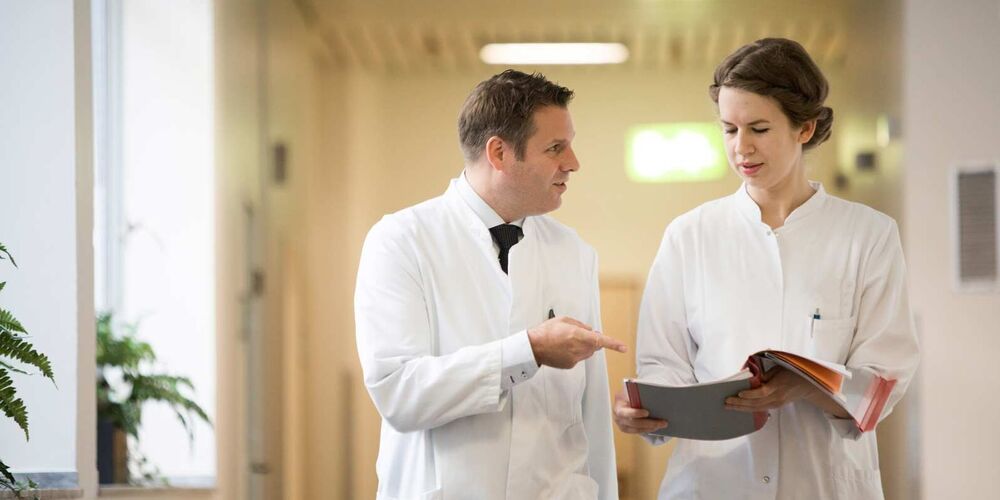 Ein Arzt und eine Ärztin unterhalten sich während sie durch den Flur eines Krankenhauses gehen. Die Ärztin hält hierbei eine offene Patientenakte in der Hand. 