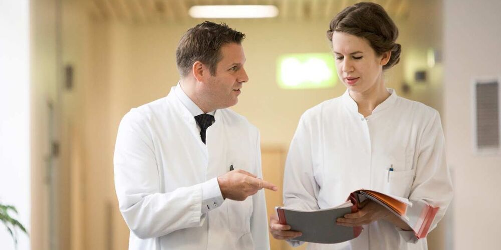 Ein Arzt und eine Ärztin unterhalten sich während sie durch den Flur eines Krankenhauses gehen. Die Ärztin hält hierbei eine offene Patientenakte in der Hand. 