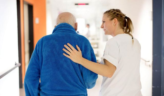 Pflegerin begleitet Senioren durch den Flur und hat ihm eine Hand auf den Rücken.