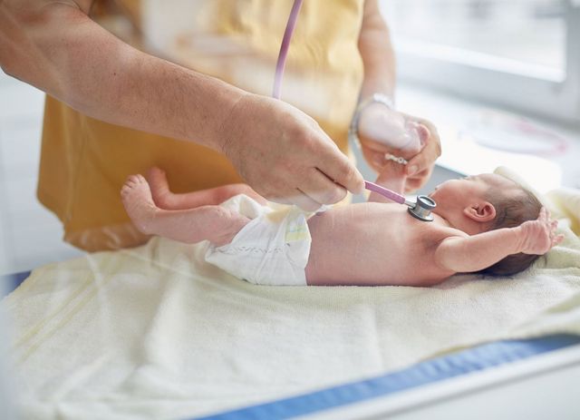 Ein Arzt untersucht ein Neugeborenes, welches nur eine Windel trägt mit einem Stethoskop auf der Brust. 